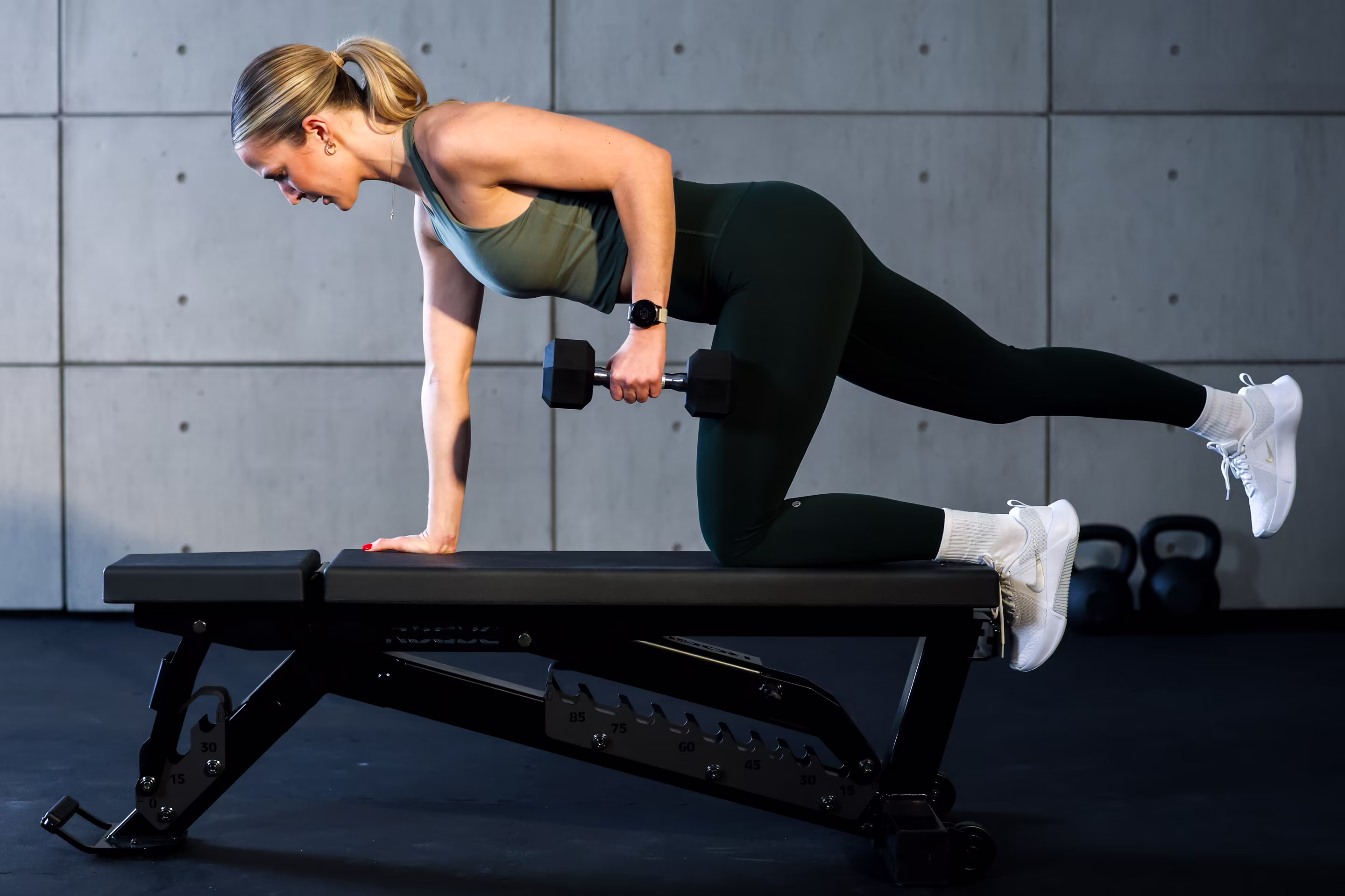 A woman rows a dumbbell with one knee on a bench