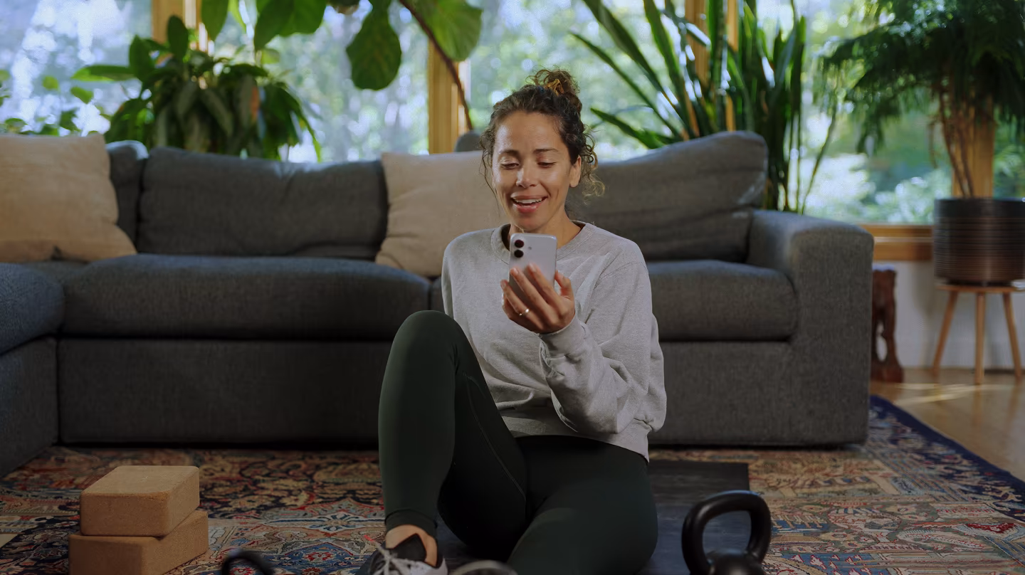 A woman sits on her living room floor and smiles at her phone