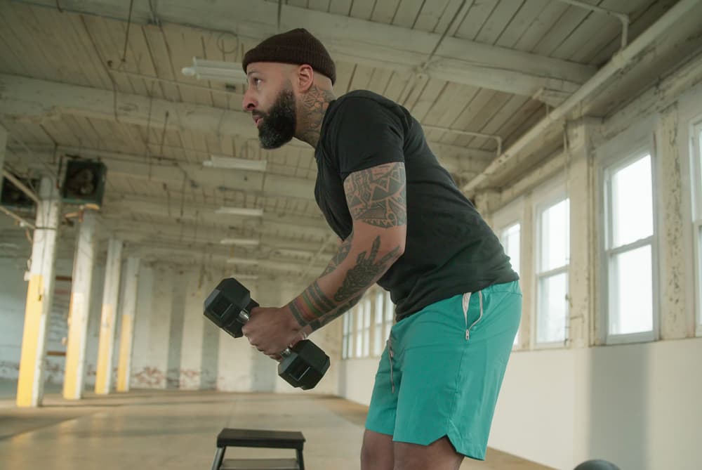 Bearded man in a beanie doing a bent-over dumbbell row in a warehouse gym