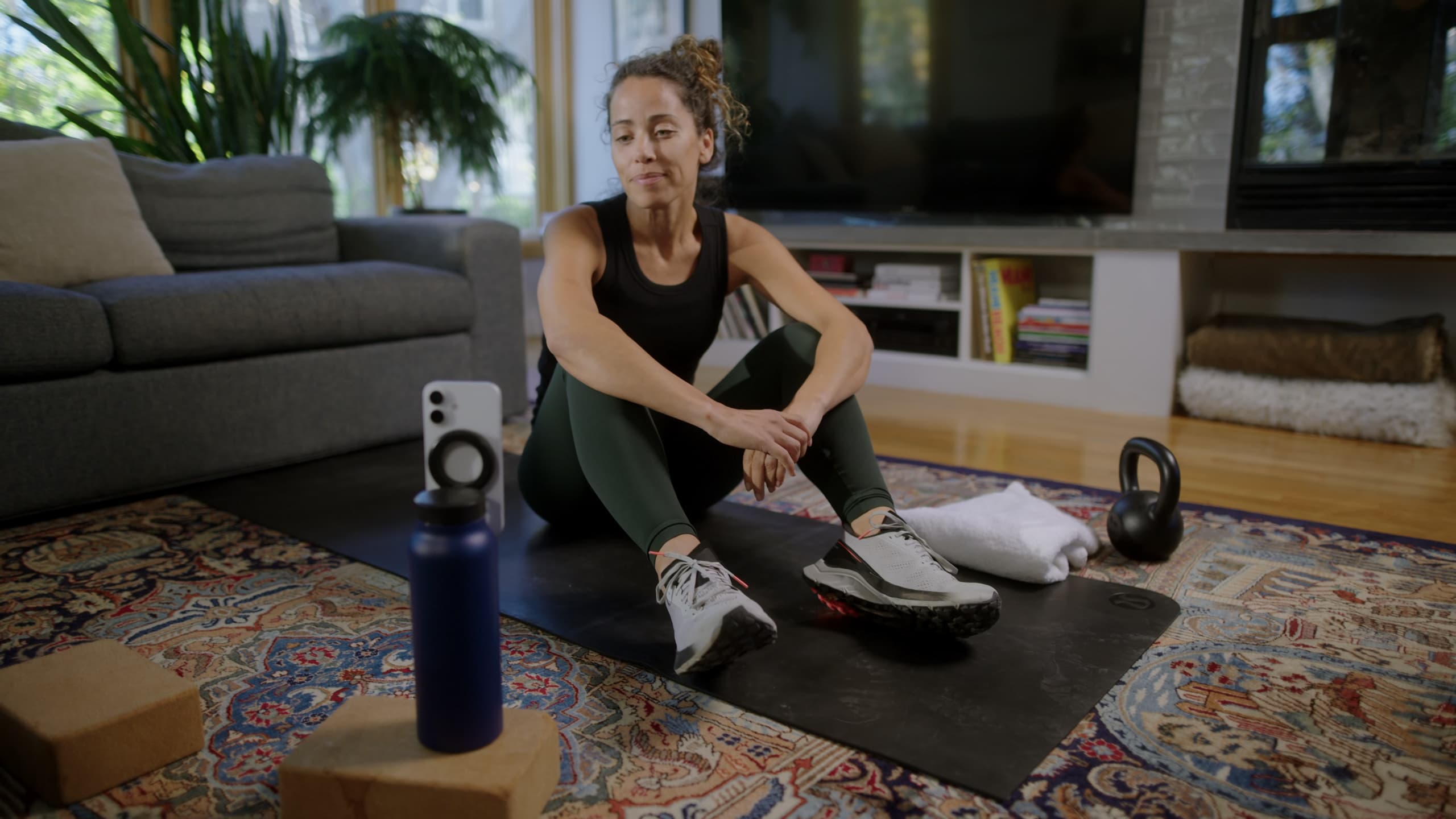 A woman sits on an exercise mat at home and smiles at her phone