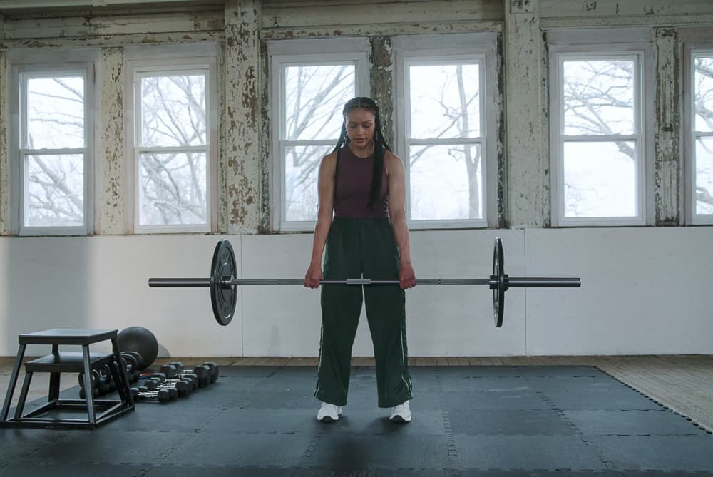 Woman with long braids standing tall at the top of a barbell deadlift in a warehouse gym