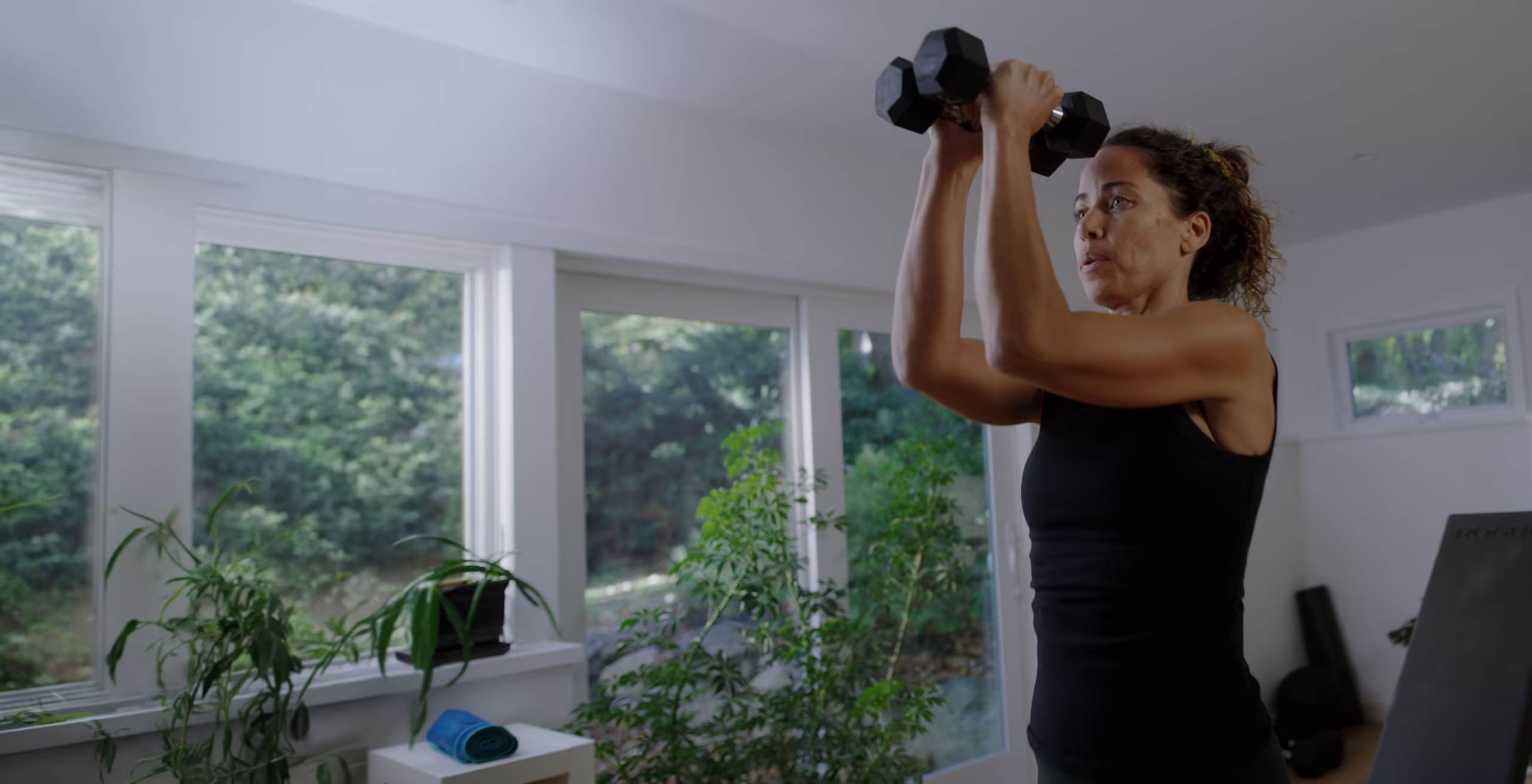 A woman presses two dumbbells overhead in a bright room with plants