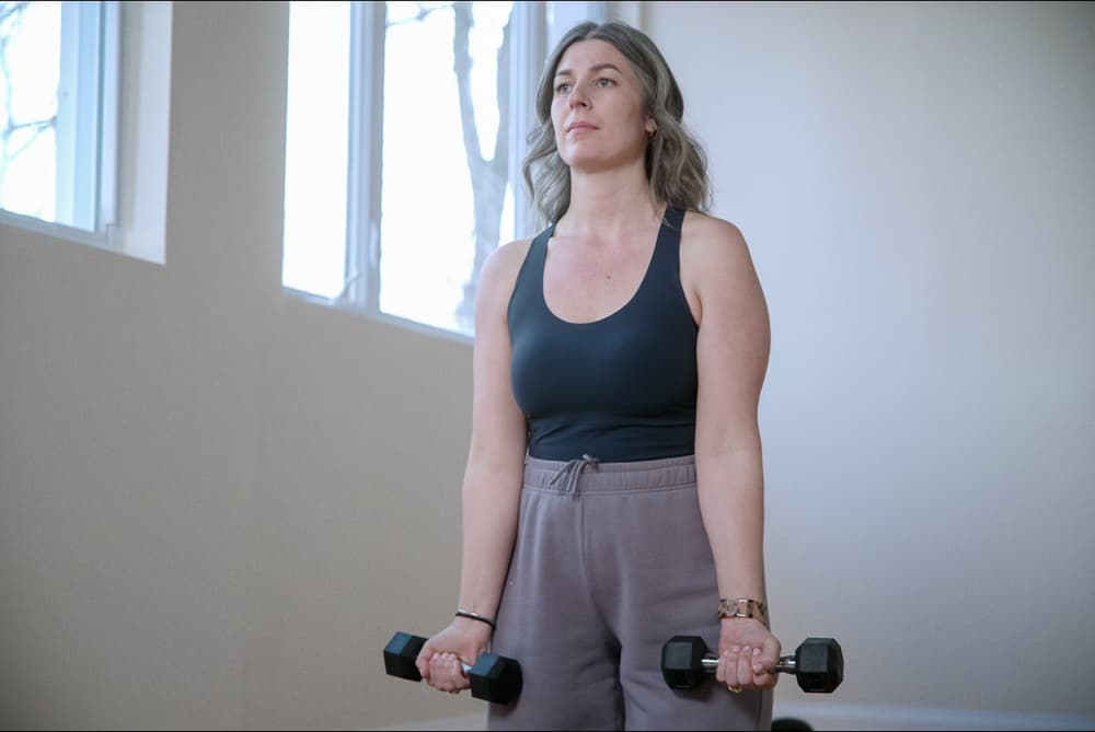 Woman with grey-streaked hair standing with a light dumbbell in each hand, ready to start a workout