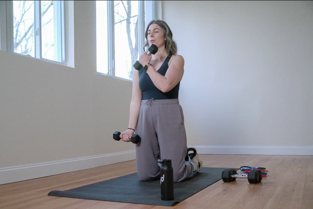 Woman kneeling on a mat doing alternating curls with light dumbbells in a bright room