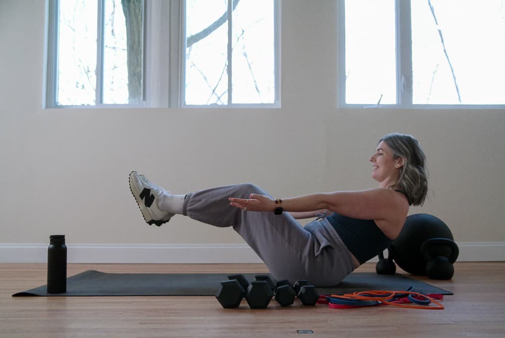 Woman smiling while holding a seated V-sit core exercise on a mat at home
